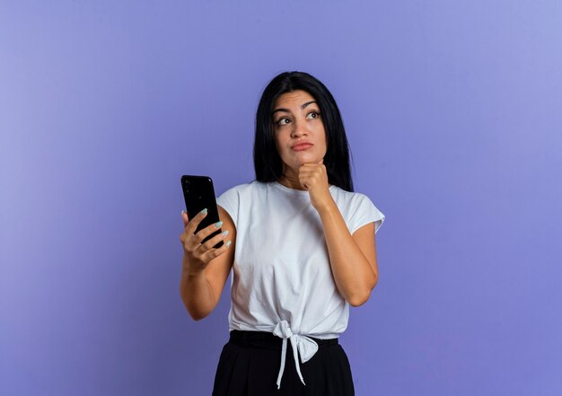 Une jeune femme caucasienne confuse tient le téléphone et met la main sur le menton en regardant de côté