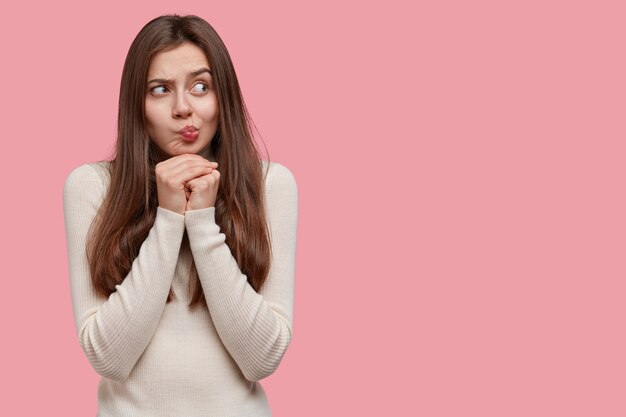 Une jeune femme caucasienne contemplative garde les mains sous le menton, fait la moue, regarde de côté avec confusion