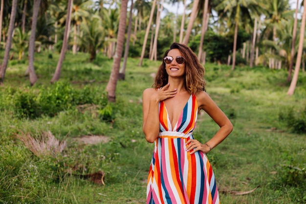 Jeune femme caucasienne heureuse en robe d'été rayée colorée avec de courts cheveux bouclés en lunettes de soleil Vacances dans un pays exotique chaud.