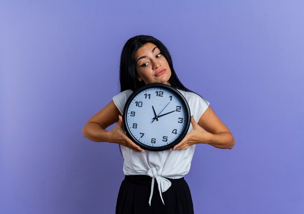 Une jeune femme caucasienne incertaine tient une horloge regardant vers le bas