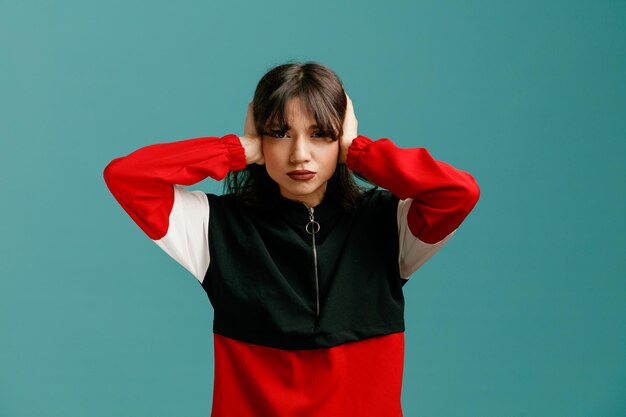 jeune femme caucasienne sérieuse regardant la caméra fermer les oreilles avec les mains isolées sur fond bleu