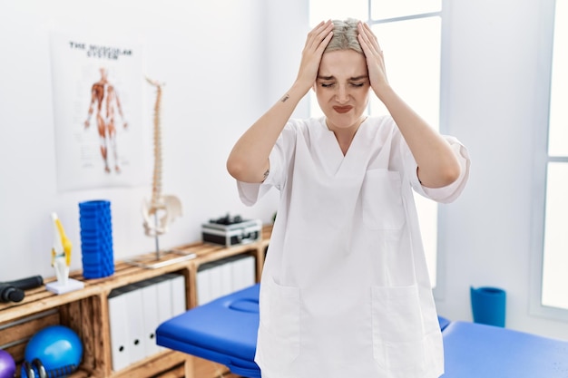 Jeune femme caucasienne travaillant à la clinique de récupération de la douleur souffrant de maux de tête désespérés et stressés à cause de la douleur et de la migraine. mains sur la tête.