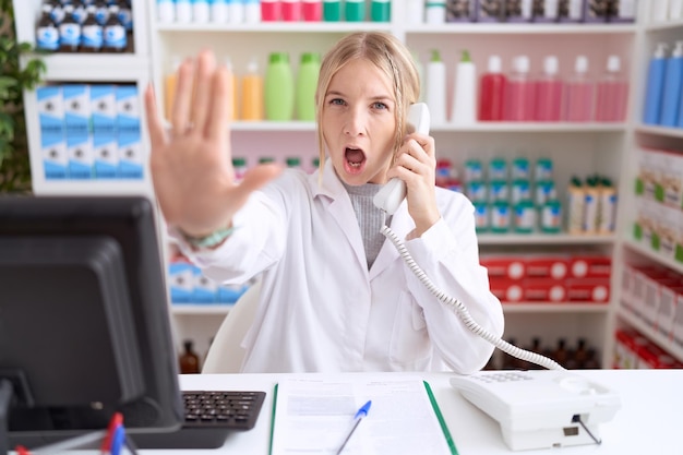 Photo gratuite jeune femme caucasienne travaillant à la pharmacie pharmacie parlant au téléphone en faisant un geste d'arrêt avec les paumes des mains en colère et expression de frustration