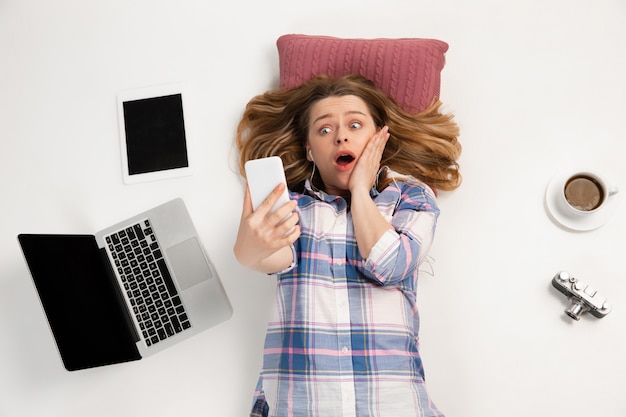 Jeune femme caucasienne utilisant des appareils, des gadgets isolés sur le mur blanc du studio.