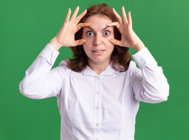 Jeune femme en chemise blanche à ar xamera ouvrant les yeux pour mieux voir debout sur mur vert