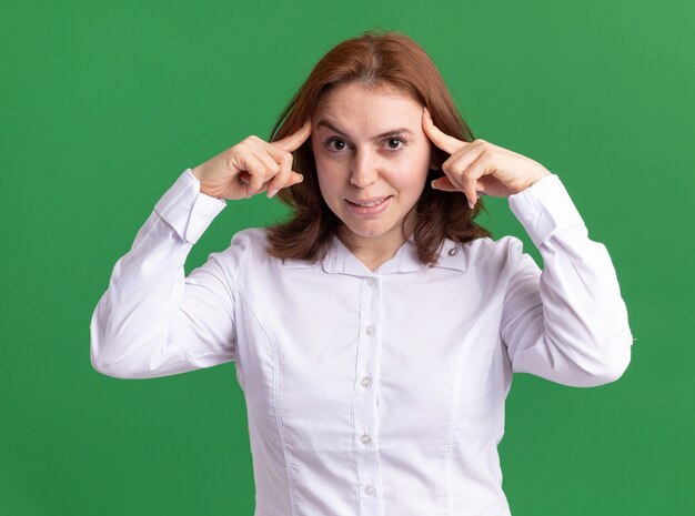 Jeune femme en chemise blanche à l'avant étant confus en pointant avec l'index sur ses tempes debout sur un mur vert