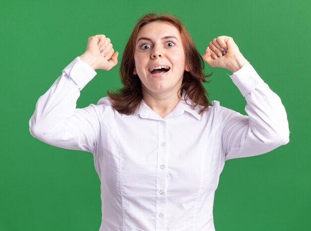 Jeune femme en chemise blanche à l'avant levant les poings heureux et excité debout sur le mur vert