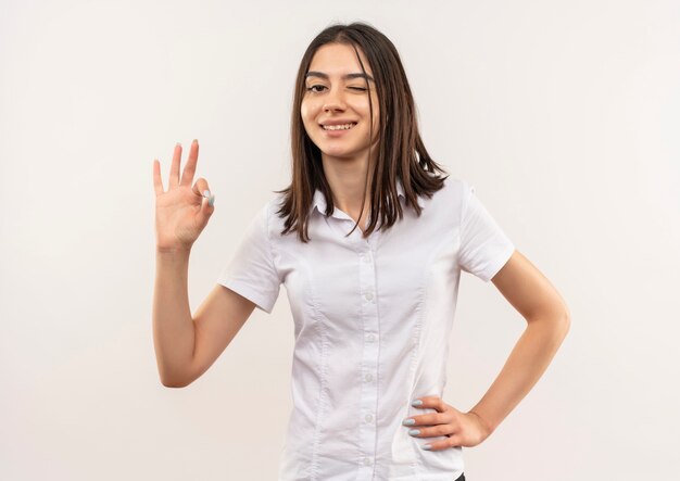 Jeune femme en chemise blanche à l'avant souriant montrant signe ok debout sur un mur blanc