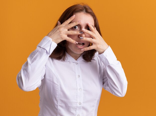 Jeune femme en chemise blanche à l'avant à travers les doigts d'avoir peur debout sur un mur orange
