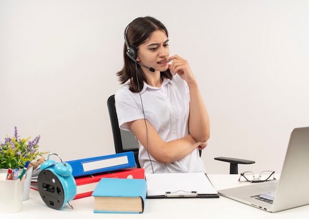 Jeune femme en chemise blanche et casque avec un microphone assis à la table avec des dossiers et un ordinateur portable en regardant son écran d'ordinateur confus sur un mur blanc