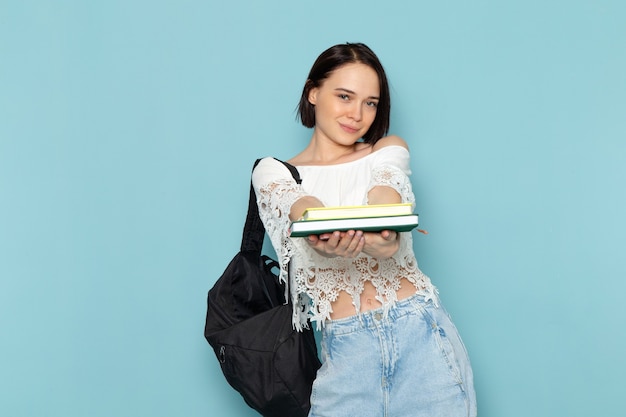 Jeune femme en chemise blanche jeans bleu et sac noir tenant des cahiers sur bleu