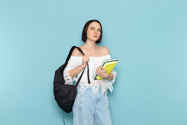 Jeune femme en chemise blanche jeans bleu et sac noir tenant des cahiers pensant sur bleu