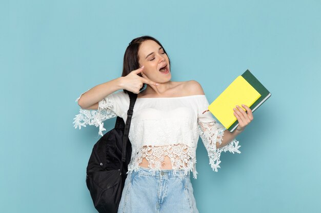 Jeune femme en chemise blanche jeans bleu et sac noir tenant des cahiers avec sourire sur bleu