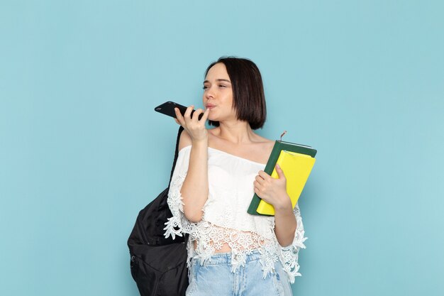 jeune femme en chemise blanche jeans bleu et sac noir tenant des cahiers et téléphone sur bleu