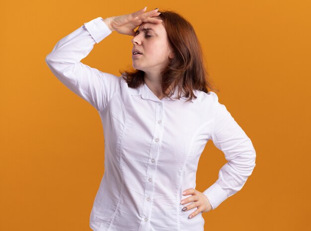 Jeune femme en chemise blanche avec la main sur sa tête fatiguée et s'ennuie debout sur un mur orange