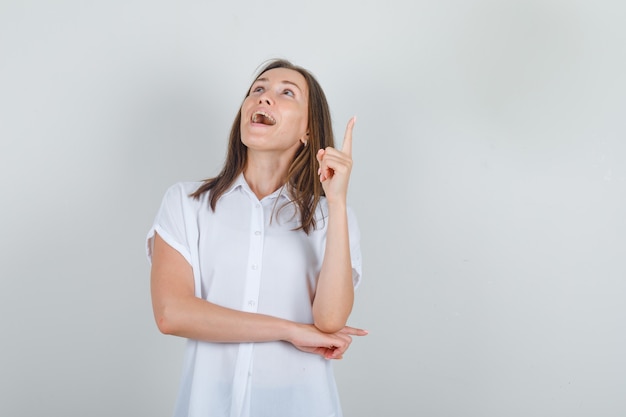 Jeune femme en chemise blanche pointant le doigt vers le haut et l'air heureux