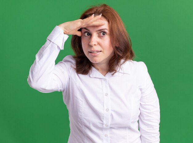 Jeune femme en chemise blanche à la recherche de loin avec la main sur la tête debout sur un mur vert