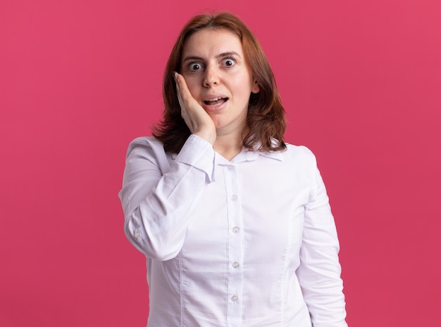 Jeune femme en chemise blanche regardant devant étonné et surpris debout sur un mur rose