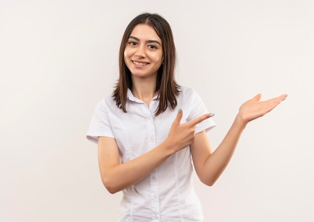 Jeune femme en chemise blanche regardant vers l'avant souriant présentant quelque chose avec le bras de sa main pointant avec le doigt sur le côté debout sur un mur blanc