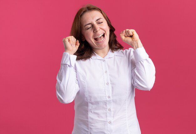 Jeune femme en chemise blanche serrant les poings heureux et excité se réjouissant debout sur le mur rose