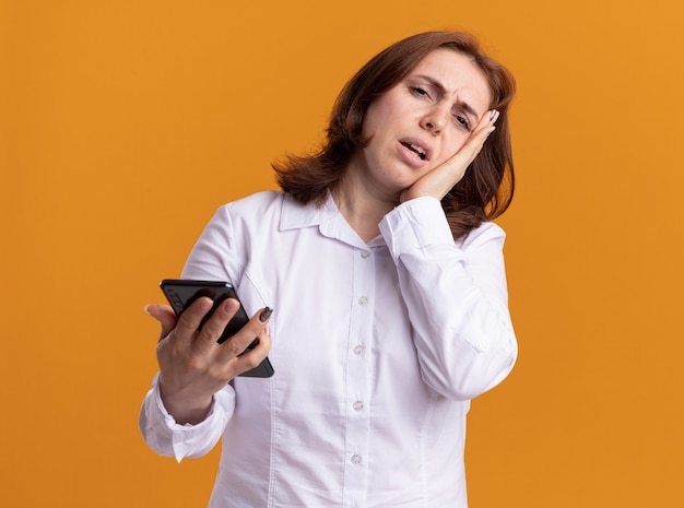 Jeune femme en chemise blanche avec smartphone à l'avant confus et mécontent debout sur un mur orange