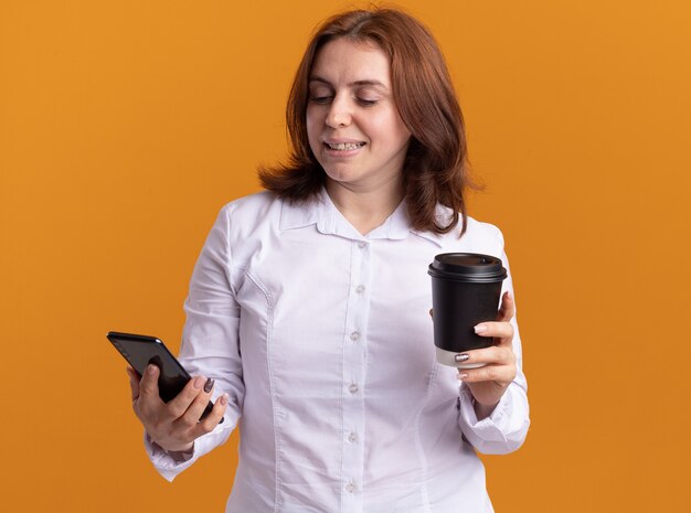 Jeune femme en chemise blanche avec smartphone tenant une tasse de café en regardant son mobile avec sourire sur le visage debout sur un mur orange