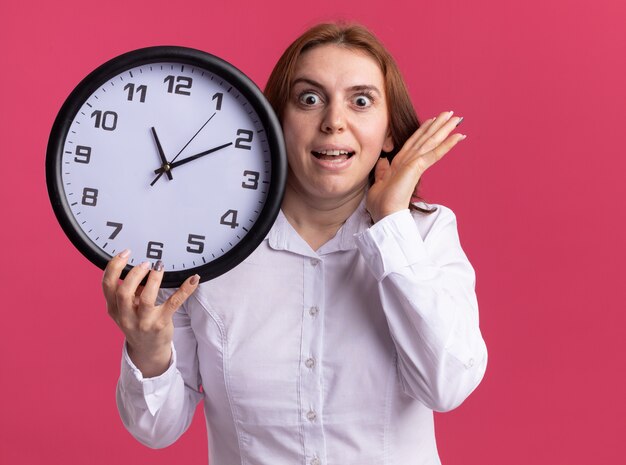 Jeune femme en chemise blanche tenant une horloge murale à l'avant étonné et surpris debout sur un mur rose