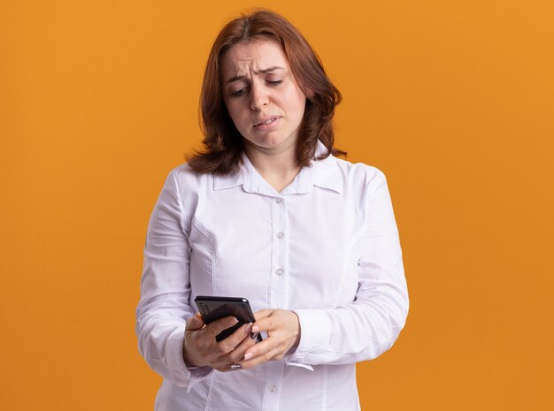 Jeune femme en chemise blanche tenant un smartphone en le regardant avec une expression triste debout sur un mur orange