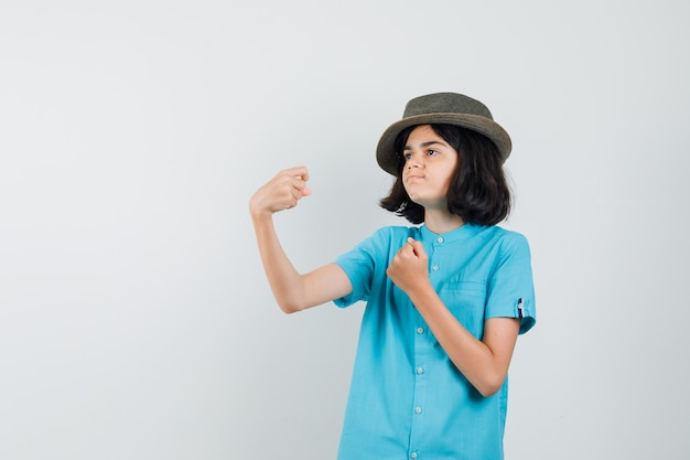 Jeune femme en chemise bleue, chapeau montrant ses muscles des bras et à la recherche d'énergie