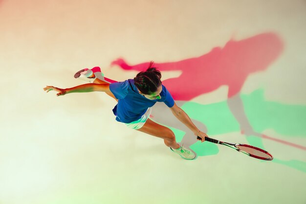 Jeune femme en chemise bleue jouant au tennis. Elle frappe la balle avec une raquette. Prise de vue en intérieur avec une lumière mixte. Jeunesse, flexibilité, puissance et énergie. Vue de dessus.