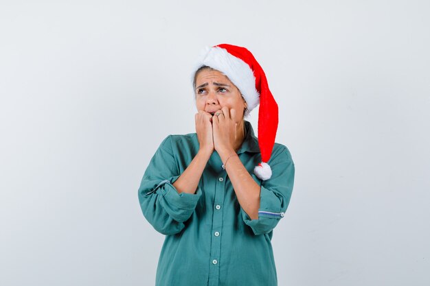 Jeune femme en chemise, bonnet de Noel gardant les mains sur le visage et l'air horrifié, vue de face.