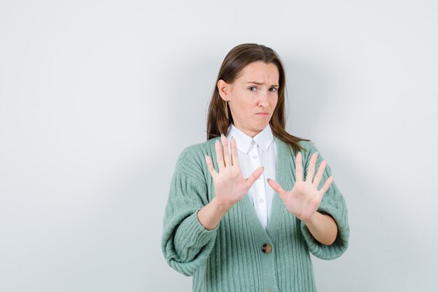 Jeune femme en chemise, cardigan montrant un geste d'arrêt et semblant sensible, vue de face.