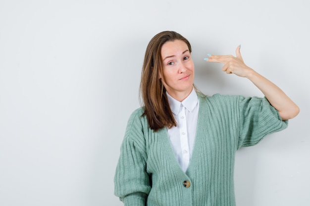 Jeune femme en chemise, cardigan montrant le geste du pistolet et l'air confiant, vue de face.