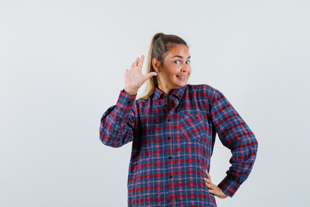 Jeune femme en chemise à carreaux agitant la main pour saluer et à la vue de face, heureux.