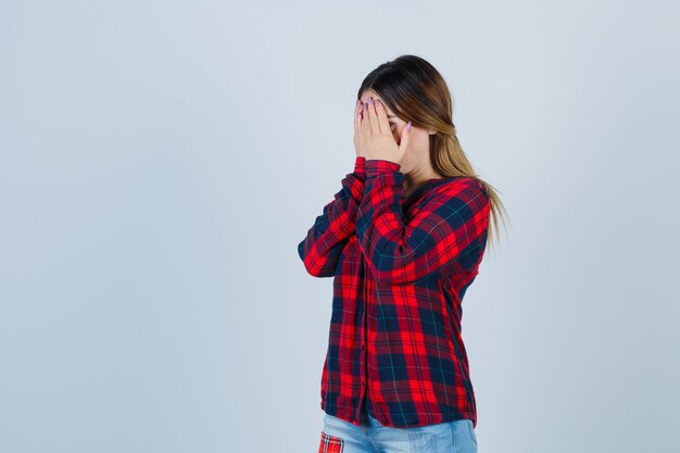 Jeune femme en chemise à carreaux couvrant le visage avec les mains et l'air déprimé, vue de face.