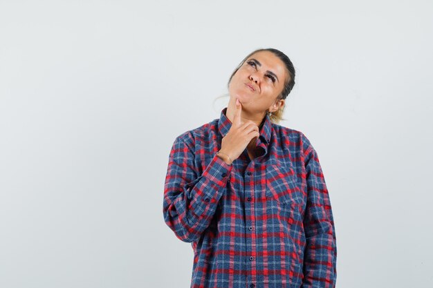 Jeune femme en chemise à carreaux debout dans la pose de la pensée et regardant vers le haut et à la vue pensive, avant