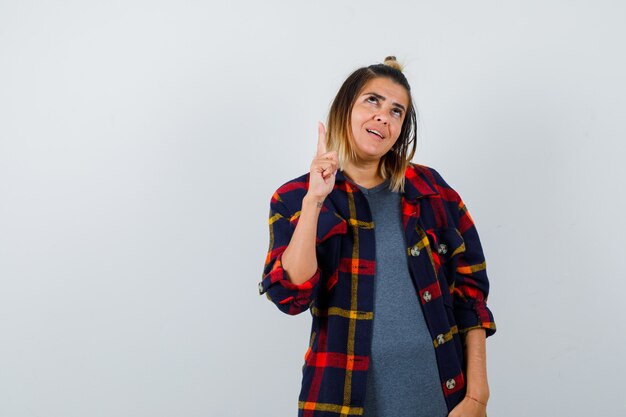 Jeune femme en chemise à carreaux décontractée pointant vers le haut et semblant attrayante, vue de face.