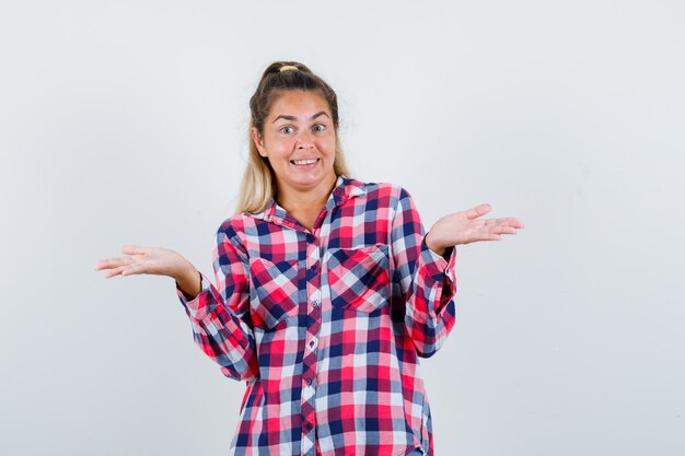 Jeune femme en chemise à carreaux étalant les paumes de côté et à la vue de face, heureux.