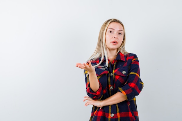 Jeune femme en chemise à carreaux, étirant la main dans le geste de questionnement et à la vue impuissante, de face.