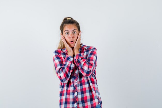 Jeune femme en chemise à carreaux gardant les mains sur les joues et à la surprise, vue de face.