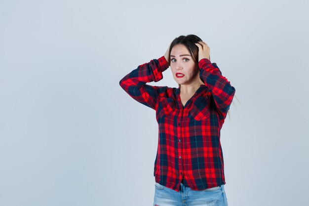 Jeune femme en chemise à carreaux, jeans avec les mains sur les oreilles et l'air effrayé, vue de face.