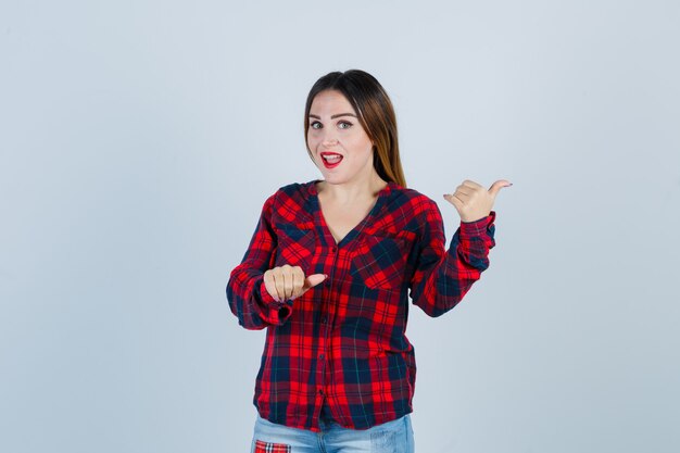Jeune femme en chemise à carreaux, jeans pointant de côté avec les pouces et l'air surpris, vue de face.