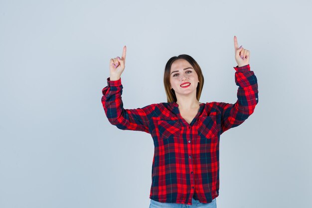 Jeune femme en chemise à carreaux, jeans pointant vers le haut en souriant et l'air heureux, vue de face.