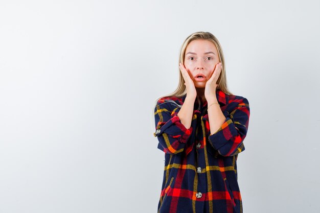 Jeune femme en chemise à carreaux, main dans la main sur les joues et à la perplexité, vue de face.