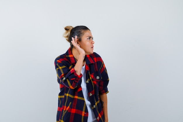 Jeune femme en chemise à carreaux avec la main derrière l'oreille, debout sur le côté et l'air curieux.