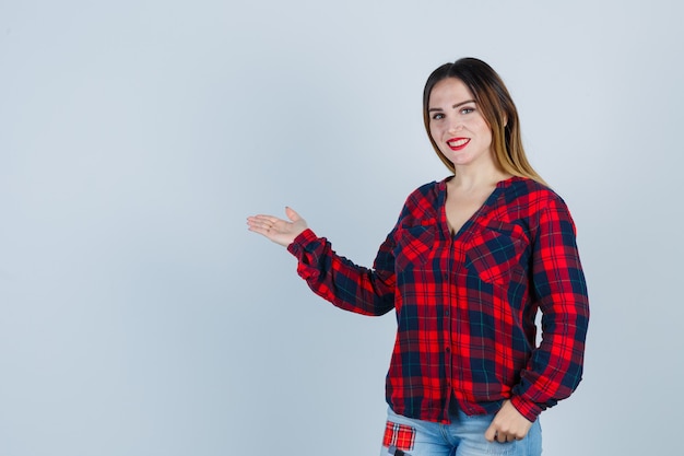 Jeune femme en chemise à carreaux montrant un geste de bienvenue et l'air confiant, vue de face.