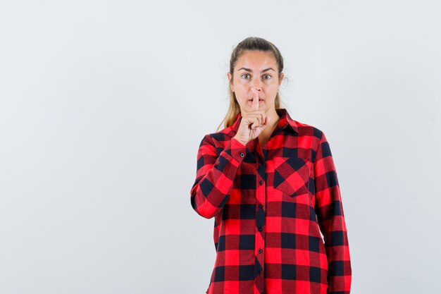 Jeune femme en chemise à carreaux montrant le geste de silence et à la prudence