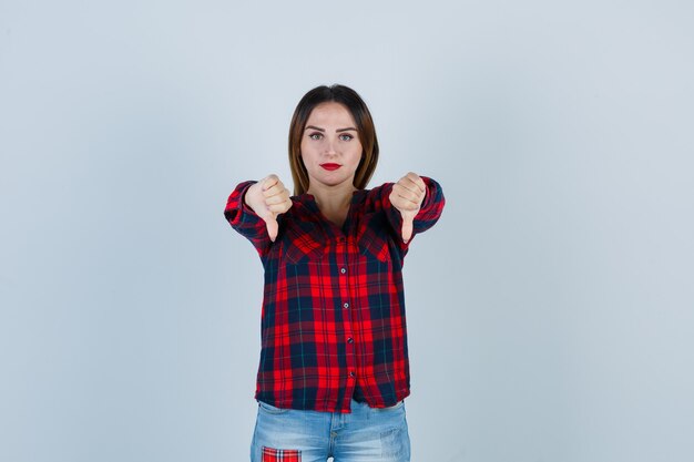 Jeune femme en chemise à carreaux montrant les pouces vers le bas et l'air confiant, vue de face.