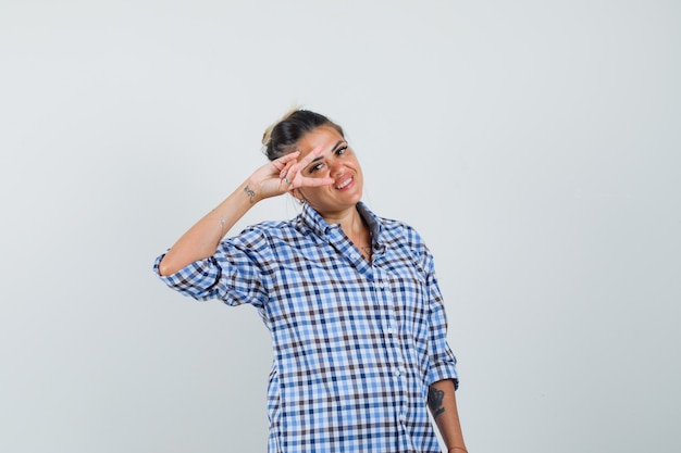 Jeune femme en chemise à carreaux montrant le signe v sur les yeux et à la joyeuse.