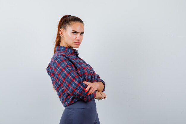 Jeune femme en chemise à carreaux, pantalon debout avec les bras croisés et l'air confiant, vue de face.